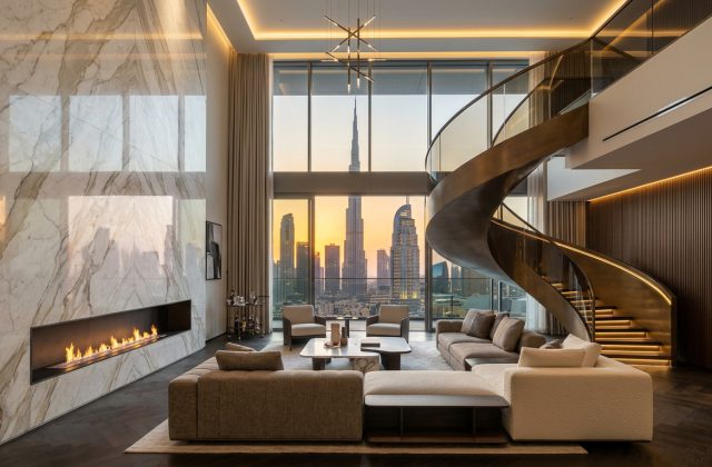 Penthouse interior design Dubai ultra-luxury double-height living salon with 7-meter panoramic glass facade overlooking Burj Khalifa skyline, bookmatched Calacatta Oro marble fireplace, and sculptural bronze staircase